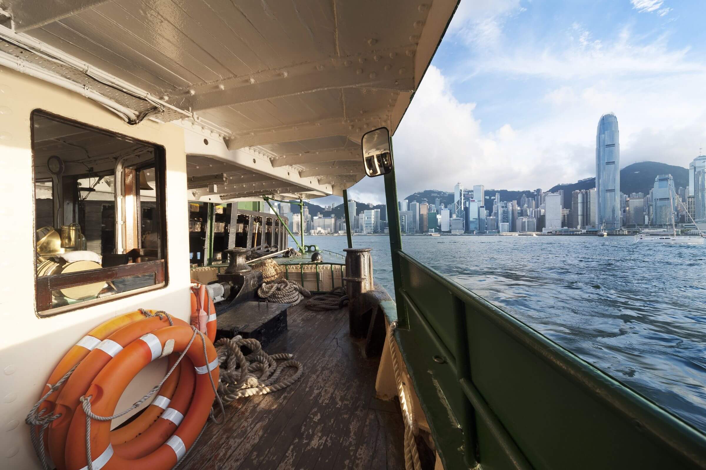 A boat is sailing across Victoria Harbour towards the Hong Kong skyline, with the city’s skyscrapers, including the International Finance Centre, visible in the background under a partly cloudy sky.